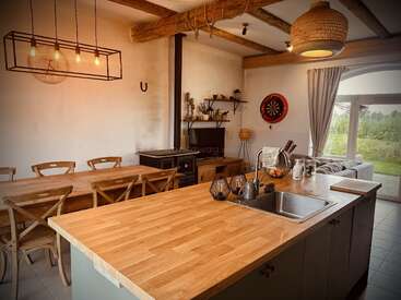 This cozy kitchen and dining area features rustic wooden beams, a large table, open shelves, modern lighting, a dartboard, and an inviting view of greenery.