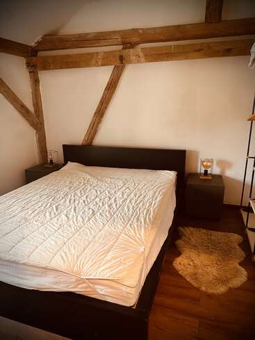 This cozy bedroom features a neatly made bed with a white comforter, wooden beams, two nightstands with lamps, and a fluffy beige rug on wooden flooring.
