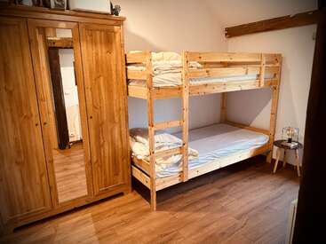 This cozy bedroom features a wooden bunk bed with white bedding, a large wooden wardrobe with a mirror, and a small round side table with a lamp.