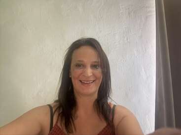 A woman with long dark hair is smiling at the camera. She stands in front of a textured white wall, wearing a sleeveless top. Natural lighting.