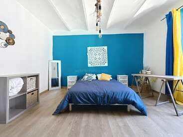 A modern bedroom with a blue accent wall, double bed, geometric artwork, natural wood floors, desk, mirror, shelving, and bright yellow and blue curtains.