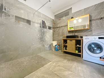 This image shows a modern bathroom with grey tiles, a glass shower, a sink with a wooden cabinet, a washing machine, and a mirror above the sink.