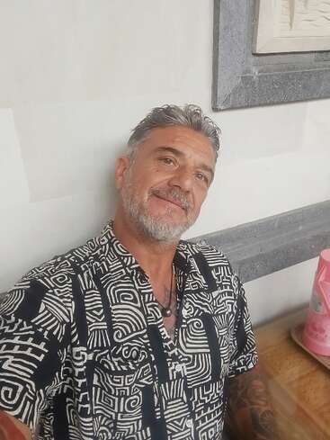 Un hombre de mediana edad con pelo y barba grises sonríe a la cámara. Lleva una camisa estampada, está sentado en el interior y a su lado se ve un recipiente rosa. Ambiente relajado.