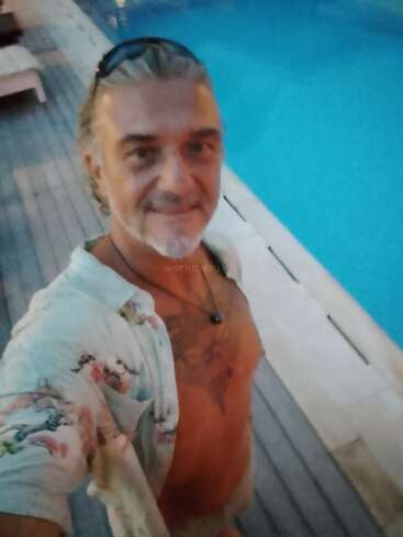 Un hombre sonriente con el pelo canoso se toma un selfie junto a una piscina azul, con una camisa de flores abierta y gafas de sol en la cabeza. El entorno es relajante.