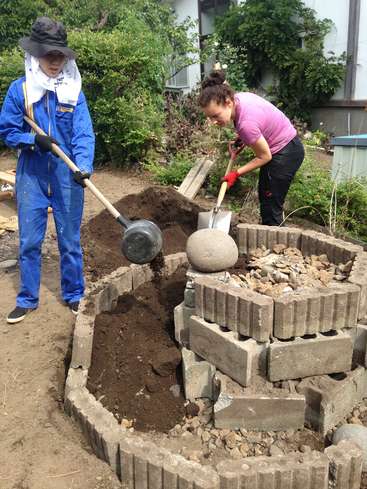 Two people are working on a garden project, using shovels to fill a circular pit with dirt, surrounded by bricks and rocks.