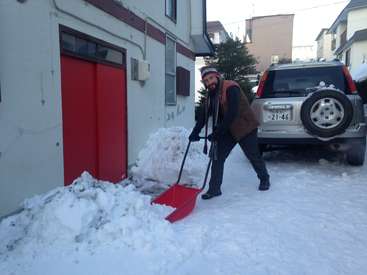A man is shoveling snow in front of a building with a red door, wearing a brown vest and hat, with a silver SUV parked nearby.