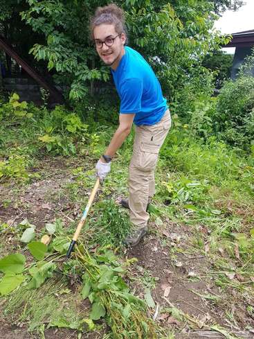 A man with glasses and a blue shirt is raking leaves and weeds in a garden, wearing gloves and standing in a yard with trees and bushes.