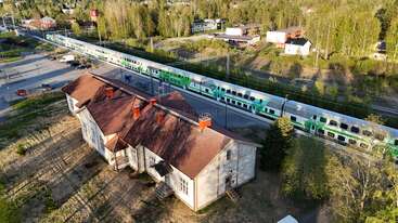 Una vista aérea muestra un edificio antiguo y grande de madera con tejado rojo, junto a un moderno tren verde y blanco que atraviesa una zona frondosa y semirural.