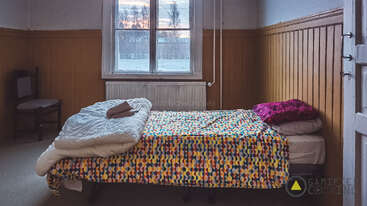 Un acogedor dormitorio con paredes revestidas de madera, una colorida colcha de lunares, la ropa de cama cuidadosamente doblada, una ventana con vistas a unos árboles nevados, una silla y unos cojines apilados junto a la cama.