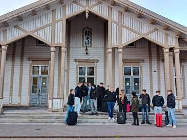 Un grupo de personas con maletas está de pie charlando frente a un edificio de madera con grandes ventanas y columnas, lo que sugiere que están esperando o a punto de viajar a algún lugar.