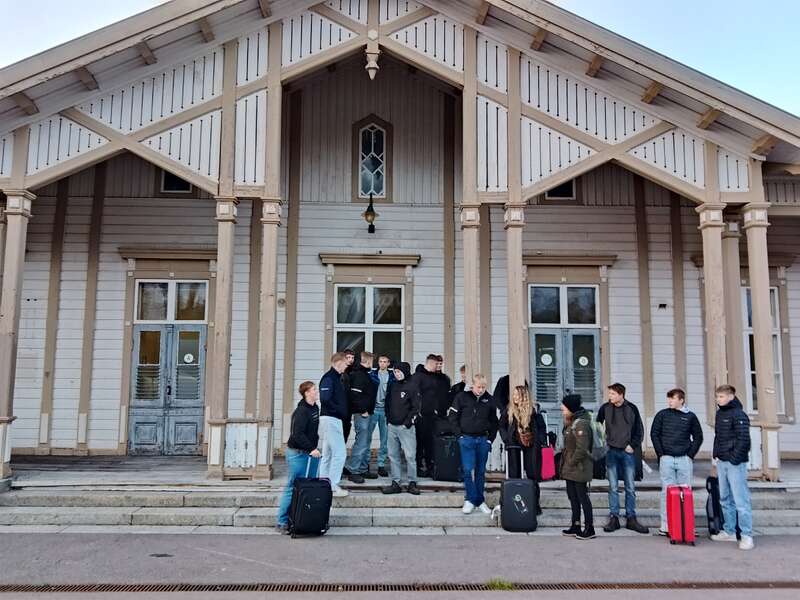 Un grupo de personas con maletas está de pie charlando frente a un edificio de madera con grandes ventanas y columnas, lo que sugiere que están esperando o a punto de viajar a algún lugar.