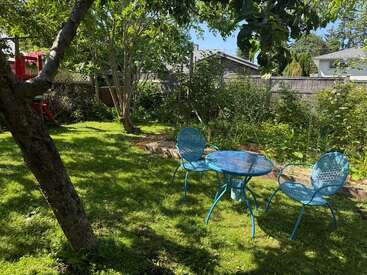 Um quintal ensolarado com grama verde, árvores e plantas. Três cadeiras de metal azuis e uma mesa combinando ficam na sombra, proporcionando uma área de estar tranquila.