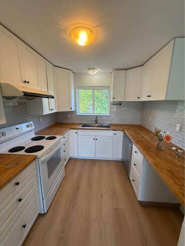 Essa cozinha tem armários brancos, bancadas de madeira, piso de madeira clara, fogão elétrico branco, máquina de lavar louça, backsplash de azulejos de metrô, janela acima da pia e flores decorativas.