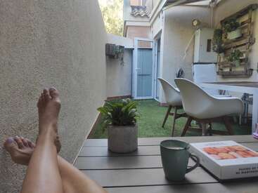 Das Bild zeigt eine ruhige Terrassenszene im Freien mit den Beinen eines Menschen, einem Tisch mit einer Pflanze und einem Becher sowie einem Essbereich vor dem Hintergrund eines Hauses und üppigem Grün.