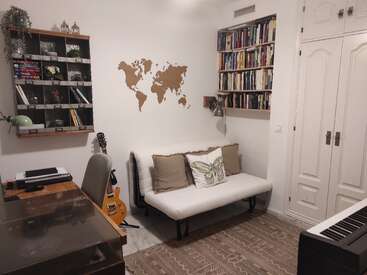 Das Bild zeigt ein gemütliches Zimmer mit einer weißen Couch, Bücherregalen, einem Schreibtisch und einem Klavier, mit einer Weltkarte an der Wand und einer Gitarre, die an der Couch lehnt.