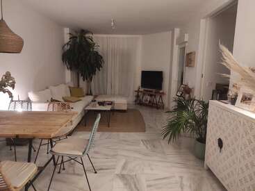 Das Bild zeigt ein modernes Wohnzimmer mit weißen Wänden, Marmorböden und einer minimalistischen Ästhetik mit einem Esstisch, einer Couch, einem Fernseher und Pflanzen.