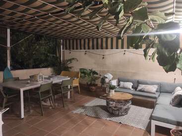 Das Bild zeigt eine gemütliche Terrasse mit einer gestreiften Markise, auf der eine Couch, ein Couchtisch, ein Esstisch und Stühle stehen, umgeben von Pflanzen und einer warmen Atmosphäre.