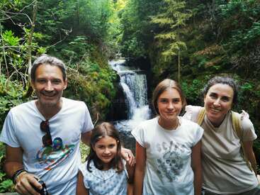 Das Bild zeigt eine vierköpfige Familie, die vor einem Wasserfall inmitten von üppigem Grün posiert, wobei der Vater und die beiden Töchter links zusammen stehen.