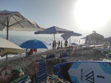 Das Bild zeigt eine ruhige Strandszene mit einem blau-weißen Paddelbrett, Sonnenschirmen, Liegestühlen und Menschen, die an einem sonnigen Tag den Blick aufs Meer genießen.