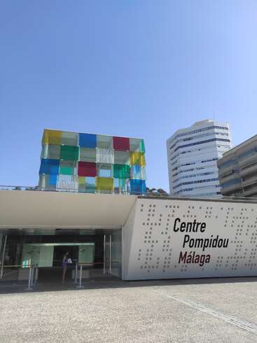 Das Bild zeigt das Centre Pompidou Málaga, ein modernes Gebäude mit einer weißen Fassade und einer lebendigen Glasstruktur vor einem klaren blauen Himmel.