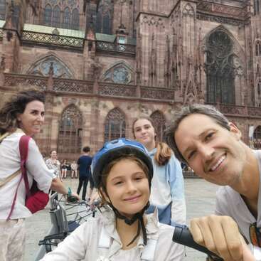 Das Bild zeigt eine vierköpfige Familie, die für ein Selfie vor einem großen Steingebäude posiert, während der Vater einen Fahrradlenker hält.