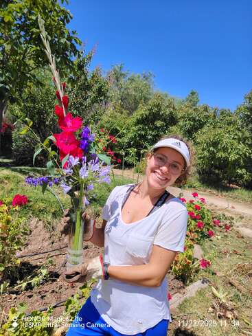 A smiling woman in a white shirt and cap holds a vase with colorful flowers in a sunny garden, surrounded by greenery and vibrant red blossoms.