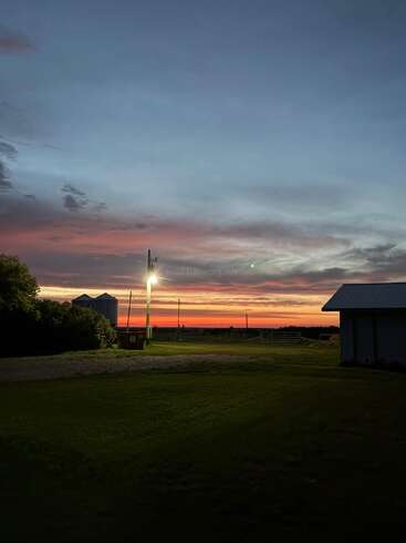 La photo capture une scène rurale tranquille au coucher du soleil, avec un ciel lumineux, des silhouettes de bâtiments, des silos, de l'herbe verte et un seul lampadaire lumineux.