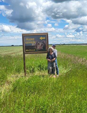 Un couple se tient dans un champ vert à côté d'un panneau "Prairie Kennels" avec une image de chien, sous un ciel partiellement nuageux. Atmosphère rurale, paisible et accueillante.