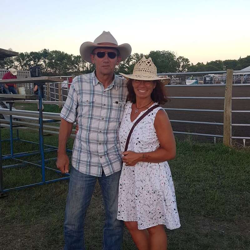 Un homme et une femme portant des chapeaux de cow-boy se tiennent ensemble et sourient lors d'un rodéo. Ils sont habillés de façon décontractée avec des clôtures, de l'herbe, des gradins et une arène à l'arrière-plan.