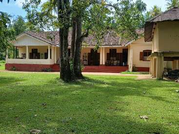 A traditional single-story house with a tiled roof stands amid lush greenery, surrounded by trees and an expansive lawn, exuding a peaceful, rural atmosphere.