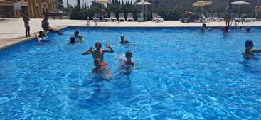 Un grupo de niños y adultos disfruta de un día soleado nadando en una piscina al aire libre de aguas cristalinas. En la zona del borde de la piscina, al fondo, hay tumbonas y sombrillas.