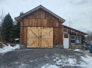 A rustic wooden barn with large double doors stands on a snowy, gravel driveway, surrounded by trees and cloudy skies, with a weathered side entrance.