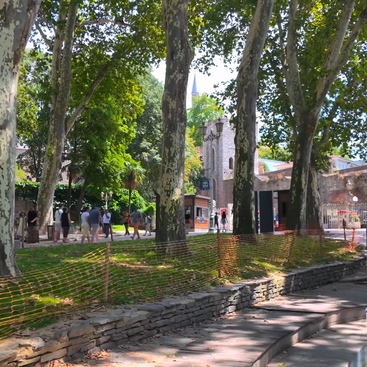 Tall trees line a sunny pathway, where people stroll near a historic stone building. Orange fencing borders a grassy area, creating a peaceful, shaded scene.