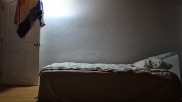 A dimly lit room features a simple single bed with white bedding and a pillow. A towel hangs on a door; the wall is plain and undecorated.