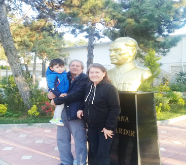 Eine dreiköpfige Familie posiert im Freien neben einer goldenen Büstenstatue. Der Mann hält ein Kind, und Bäume und Grünpflanzen umgeben die gepflasterte Fläche und die Statue.