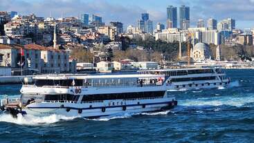 Zwei Fähren fahren über das blaue Wasser mit den Wellen, vor einer pulsierenden Stadtsilhouette mit modernen Wolkenkratzern, historischen Gebäuden und einer Moschee an Land.
