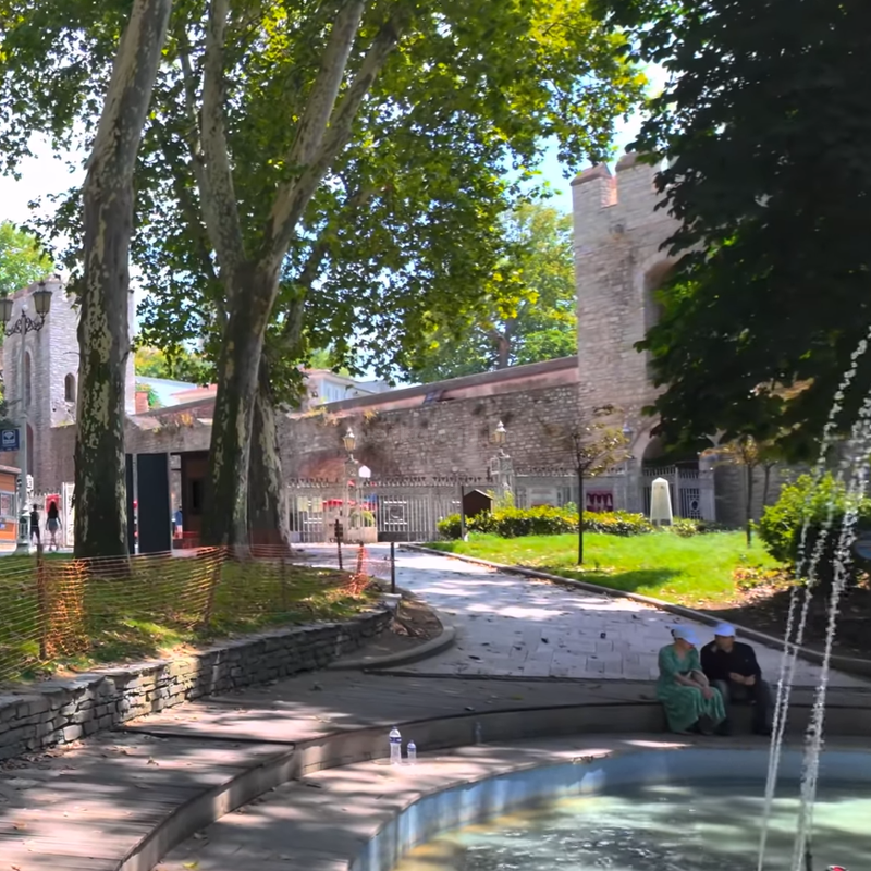 Tall trees line a sunny pathway, where people stroll near a historic stone building. Orange fencing borders a grassy area, creating a peaceful, shaded scene.