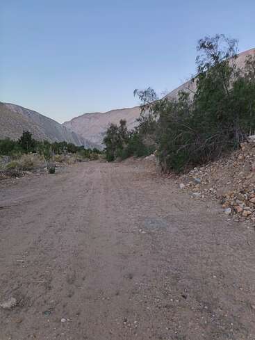 Eine unbefestigte Straße führt durch ein zerklüftetes, trockenes Tal, flankiert von felsigen Hügeln und verstreuten Büschen, unter einem klaren blauen Himmel in der Abenddämmerung. Friedliche Szenerie.