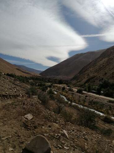 Eine felsige, karge Landschaft mit verstreuten Sträuchern und einem gewundenen Fluss. Majestätische Berge erheben sich unter dramatischen, strukturierten Wolken in einem leuchtenden, weitläufigen Himmel. Die Natur fühlt sich hier riesig an.