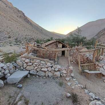 Eine rustikale Hütte aus Stein und Holz steht bei Sonnenuntergang in einer trockenen Berglandschaft, umgeben von Felswänden und einfachen Holzzäunen, die an ein abgelegenes Landleben erinnern.