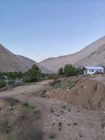 Ein ruhiges Tal mit zerklüfteten Hügeln, verstreutem Grün, einem Feldweg und einem kleinen weißen Haus mit einem roten Auto. Der Himmel ist klar und blau.