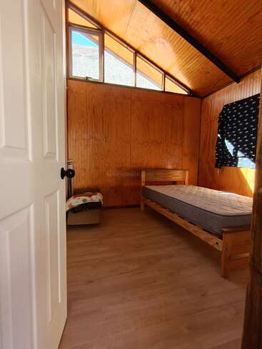 Dieses Bild zeigt ein einfaches, sonnenbeschienenes Zimmer mit Holzwänden und -decke, einem Einzelbett mit Matratze, einem kleinen Fenster, einem Holzboden und einer teilweise geöffneten weißen Tür.