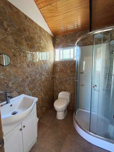 Dieses Badezimmer hat marmorierte Wände, ein weißes Waschbecken mit Unterschrank, eine Toilette, eine gebogene Glasduschkabine, einen kleinen runden Spiegel, eine hohe Holzdecke und Fenster.