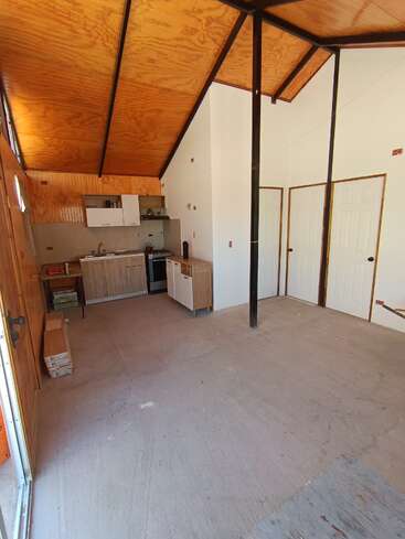 Dieses Bild zeigt einen kleinen, unfertigen Raum mit einer hohen Holzdecke, einem einfachen Küchenbereich, weißen Türen und Sichtbetonböden. Die Einrichtung ist minimalistisch.