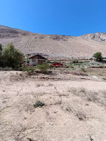 Ein kleines Haus und ein rotes Auto stehen am Fuße eines kargen, sandigen Hügels, umgeben von spärlicher Vegetation unter einem klaren, strahlend blauen Himmel.