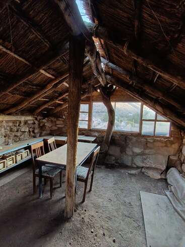 Esta imagem mostra o interior de uma cabana rústica com telhado de palha, paredes de pedra, móveis de madeira, piso de terra batida e grandes janelas que permitem a entrada de luz natural.