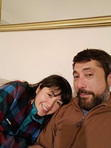 Eine Frau lehnt ihren Kopf an die Schulter eines Mannes, beide lächeln warmherzig. Sie sitzen auf einer Couch mit einem großen goldgerahmten Spiegel an der Wand hinter ihnen.