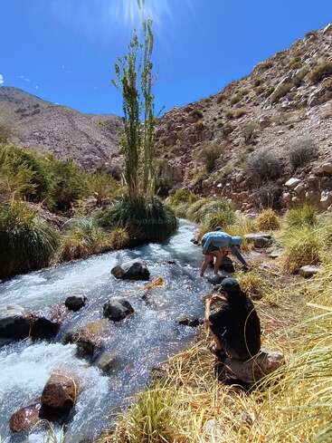 Duas pessoas desfrutam de um riacho de montanha límpido sob o sol forte, cercado por colinas rochosas, plantas verdes e um céu azul vibrante em uma paisagem natural tranquila.