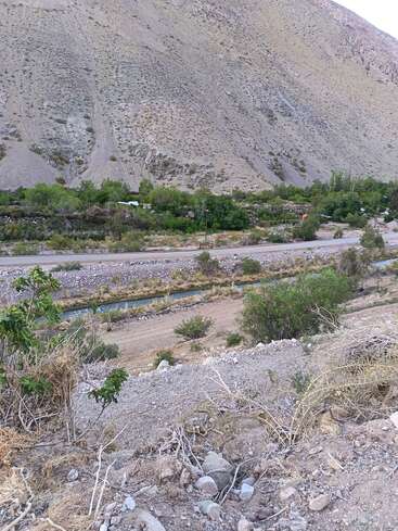 Esta imagem mostra uma encosta seca e rochosa com vegetação esparsa. Abaixo, uma estrada de terra e um pequeno canal são cercados por árvores e arbustos verdes, com montanhas.