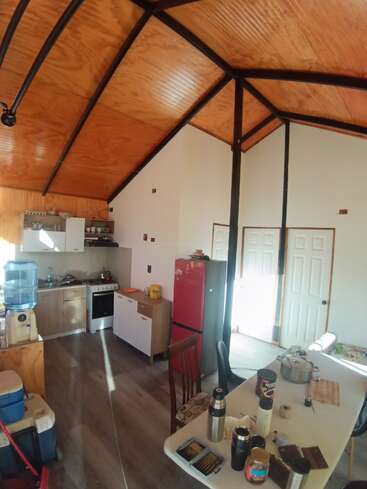 This image shows a cozy kitchen and dining area with a wooden ceiling, red fridge, white cabinets, table with food items, and two white doors in back.